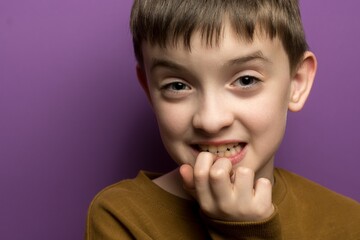A nervous boy in a brown long-sleeve shirt bites his nails, looking anxious against a purple background.