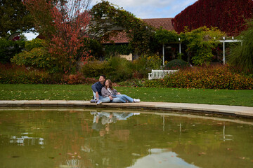 Romantic young couple relaxing by the pond in autumn park, wearing cozy casual outfits, enjoying a peaceful moment together.