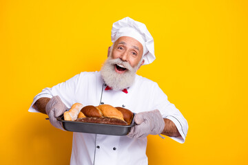 Smiling senior chef holding freshly baked bread in a baking tray with bright yellow background showcasing culinary enthusiasm