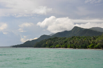 Panoramic view from the ocean to the tropical island  Karimunjawa with turquoise water and lush vegetation on the hill under a cloudy sky. Jepara regency, central Java, Indonesia