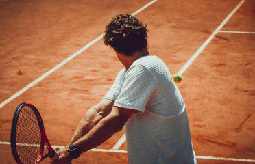 A person playing tennis on a clay court, mid-backhand swing, with the tennis ball visible near the...