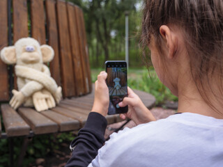 Cute girl with bun hairstyle holds smartphone and takes photo of her funny stuffed toy monkey with bulging eyes sitting on a wooden bench in a city park outdoors.