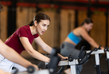 Fototapeta premium Focused girl works out with exercise bicycle during class at fitness club. Gym visitor performs exercise, creates load on muscles.