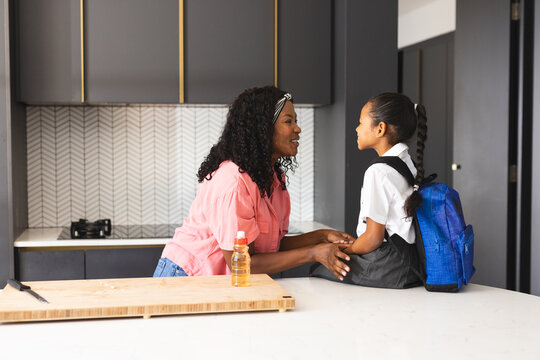 Diverse mother talking to daughter with backpack in kitchen before school, sharing smiles, at home