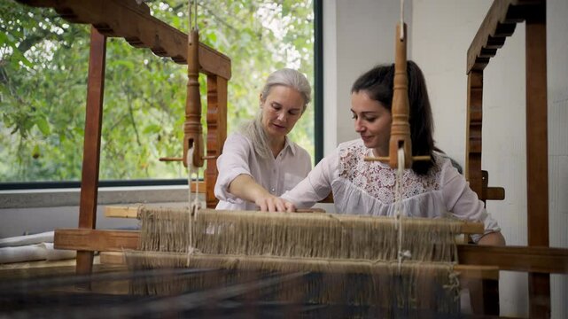 Young businesswoman with her older colleage sitting at a wooden loom, weaving a rug from old fabrics. Upcycling in business and circular economy.