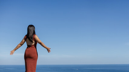 Photo of a beautiful young woman from her back, breathing and meditating, looking towards the sea and the blue sky in the background.