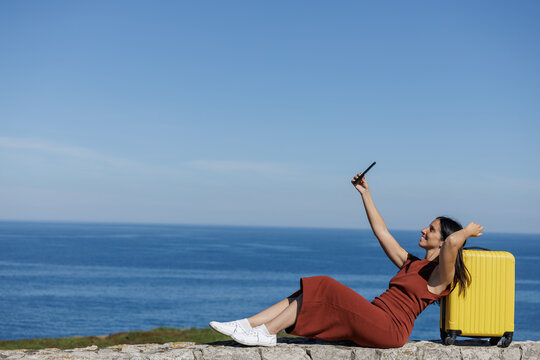 Smiling woman takes photos and selfies with her mobile phone with her yellow suitcase and the blue sky in the background, she is ready to start her summer trip. Concept of disconnection and enjoying.