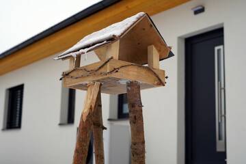 A homemade bird feeder that is handmade. It stands next to a new building in the backyard.
