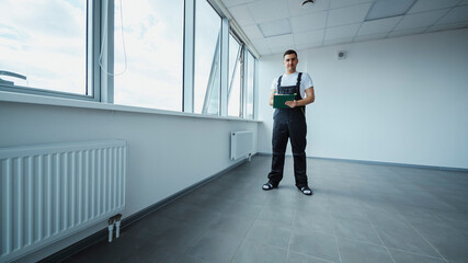 Fototapeta premium Maintenance professional checking clipboard while surveying vacant room, noting details near large windows and industrial radiator, documenting building condition carefully