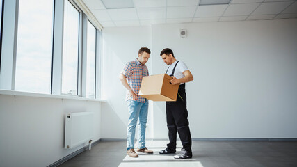 Professional movers collaborating, transporting cardboard box through spacious, sunlit office with radiator during workplace relocation