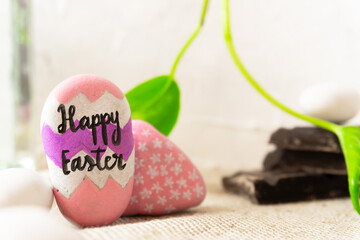 Easter background with two rocks painted by hand. One is decorated with the sentence Happy Easter. Some blocks of chocolates and vibrant green leaves are placed in the background