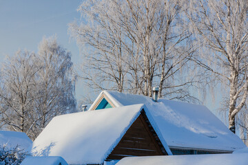 The roofs of wooden houses are covered with a thick layer of snow. In the background are tall birch trees covered with frost. Snowy winter.