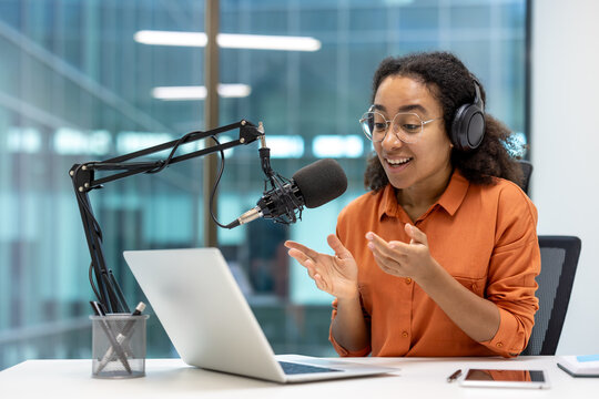 Young, cheerful woman with glasses and headphones speaks into a microphone while recording a podcast in a modern office setting