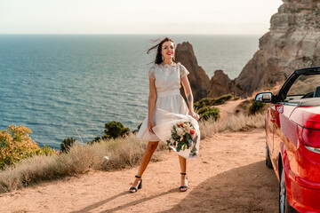 A woman in a white dress is standing next to a red convertible car. She is holding a bouquet of...