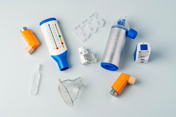 Inhalers, peak flow meter, plastic ampoules, nebulizer components, and metal spacer on white background for respiratory therapy.
