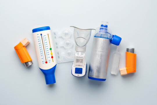 Inhalers, nebulizer mask, plastic blister pack, peak flow meter, and metal spacer on light backdrop for respiratory care.