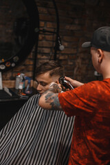 A barber man with a tattoo on his arm makes a haircut to a brunette guy in a barbershop, cuts his hair with a clipper close-up