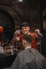 A barber man with a tattoo on his arm makes a haircut to a brunette guy in a barbershop, cuts his hair with a clipper close-up