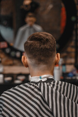 A barber man with a tattoo on his arm makes a haircut to a brunette guy in a barbershop, cuts his hair with a clipper close-up