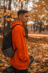 young man skater teenager in bright orange robe sitting outdoors in nature. Skateboarding in free time as a hobby. Beautiful autumn with orange leaves