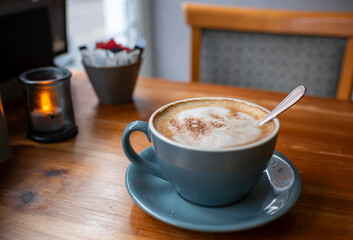 A cup of coffee sits on a saucer on a wooden table. A side shot from the viewers seat. The coffee is topped with whipped cream and a spoon is resting on the surface. The scene is cozy and inviting