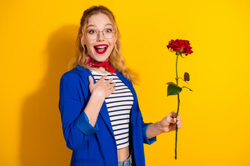 Young woman joyful over gift holding rose wearing glasses and blue jacket on a vibrant yellow background