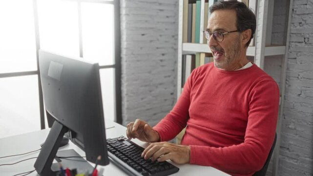 Hispanic man in red sweater works and celebrates success at computer in bright modern office setting.