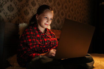 Pretty preteen girl with beautiful smile using laptop sitting in bed in dark bedroom at home, face...