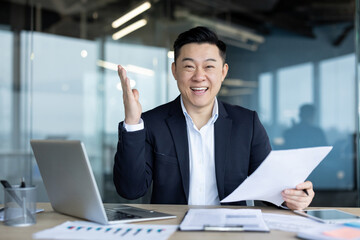 Portrait of a young happy Asian man working in the office, holding documents in his hand, satisfied with the results, looking at the camera with a smile