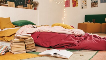 A relaxed bedroom featuring colorful bedding, stacks of books, fairy lights, and natural light streaming through wooden windows, creating a lived-in feel. Cluttercore
