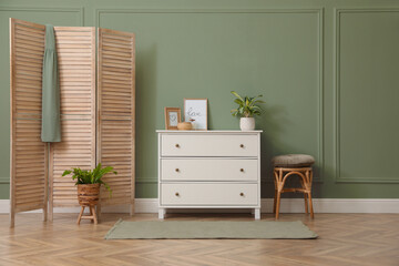 Stylish room with wooden folding screen and chest of drawers near sage color wall. Interior design