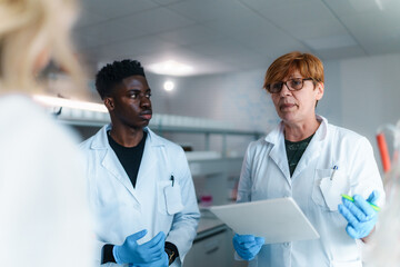 Professor holding a tablet while explaining a lesson to a diverse group of students dressed in lab coats and gloves, engaged in hands on learning in a modern university laboratory