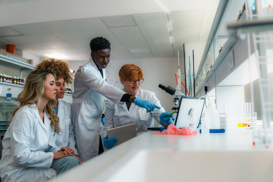 Group of diverse university students and their professor wearing lab coats and gloves are cooperating on a medical research project in a modern laboratory, using a computer and a microscope