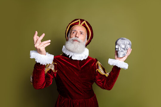 Mature man in a costume holding a skull on khaki background demonstrating historic and expressive style, think to be or not to be