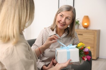 Smiling mom receiving bouquet of tulips and gift from her daughter on sofa at home. Happy Mother's...