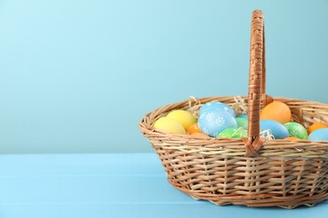 Many decorated Easter eggs in wicker basket on light blue wooden table, space for text