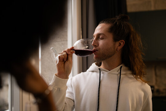Relaxed male sipping red wine by home window, closed eyes, experiencing rich flavor and fragrant notes in casual attire