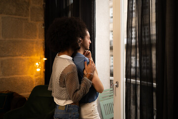 Lgbtq plus partners sharing gentle embrace near apartment window, silhouetted against urban skyline, experiencing intimate connection and quiet companionship