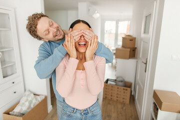 Loving man closing his girlfriend's eyes standing in their own home among cardboard boxes, guy making surprise for his beloved wife in new apartment
