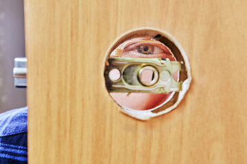 Person eye is visible through circular peephole in wooden door, with brass latch mechanism partially exposed.