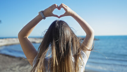 Woman forming heart with hands overlooking seaside promenade against blue sky reflecting carefree coastal atmosphere.