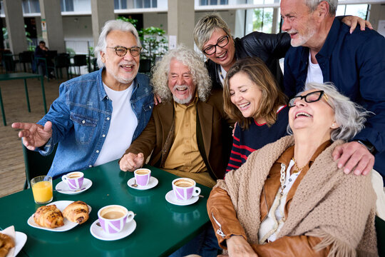 Cheerful group of mature people laughing and enjoying breakfast sitting at cafeteria bar table together. Retired generation community friends having fun gathered on terrace for snacking - Powered by Adobe