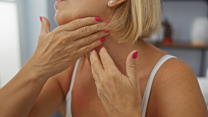 Mature woman with short blonde hair and pink nails examining her neck with hands at a salon or spa indoors, showcasing a wellness beauty treatment setting