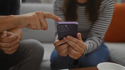 Man pointing at smartphone held by woman in living room while sitting together on sofa indoors indicating focused analysis