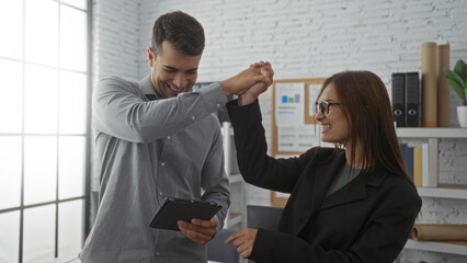 Man and woman celebrating a success together in a cozy apartment living room, both smiling and holding a tablet in a bright indoor setting, emphasizing their partnership in business