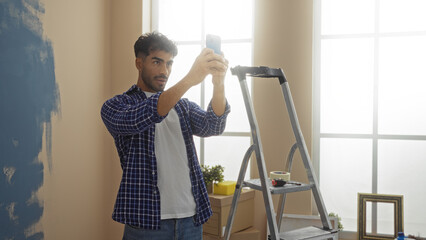 Young hispanic man taking photo with phone in newly painted living room interior apartment using ladder for decoration project during move-in day © Krakenimages.com