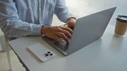 Young man working indoors on laptop in office setting with smartphone and coffee cup nearby, focused on task in casual attire indicating a professional environment.