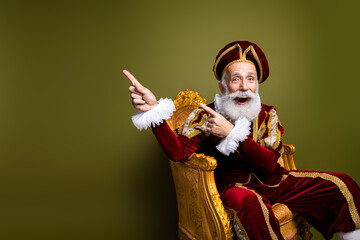 Fototapeta premium Cheerful Elderly Gentleman in Opulent Royal Costume Sitting on Ornate Chair Against Green Background