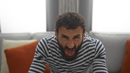 Obraz premium Young man with beard sitting in living room with striped shirt, appearing frustrated or angry, set in a cozy home setting with pillows on the sofa