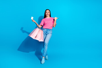 Woman holding shopping bags and credit card on blue background, wearing pink striped shirt and blue pants, expressing joy